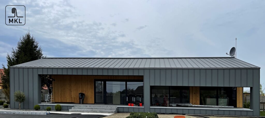 bočný pohľad na modernú budovu so zakrivenou sivou falcovanou strechou MKL, kombinácia hladkej fasády a technických prvkov, modrá obloha, presné detaily kovových spojo